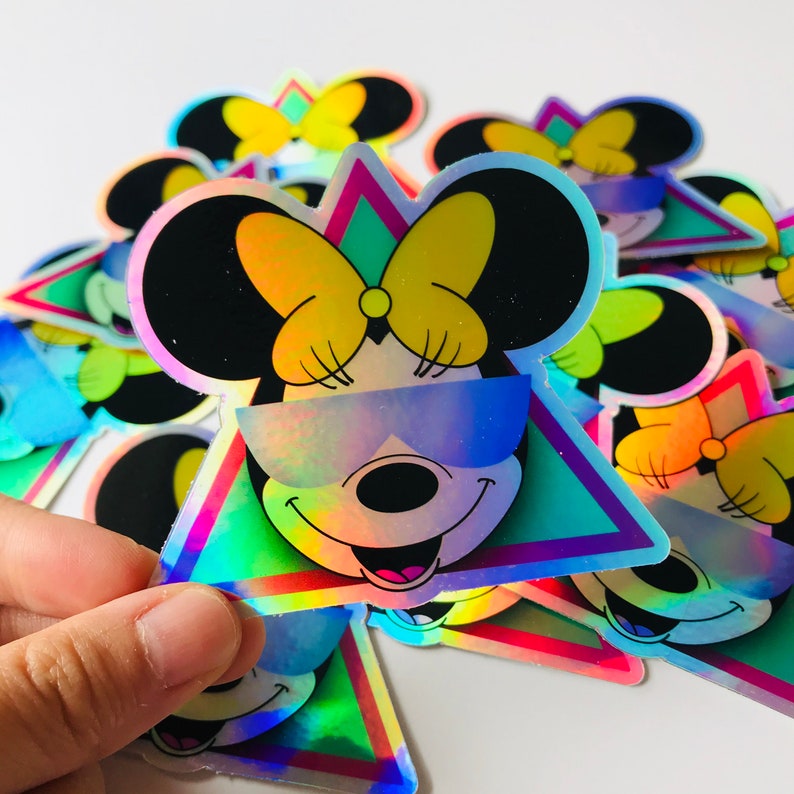 May include: Holographic sticker featuring a smiling cartoon mouse with black ears, a yellow bow, and sunglasses. The mouse is set against a teal and pink triangle. The sticker has a shiny, iridescent finish.