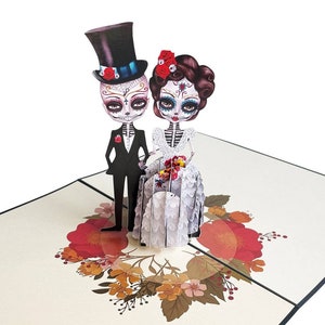 May include: A 3D pop-up card depicting a skeleton bride and groom in traditional wedding attire. The bride is wearing a white dress with floral embellishments and a red flower in her hair. The groom is wearing a black suit and a top hat. The card is adorned with a floral wreath.