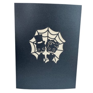 May include: A black card with a white cutout design of a couple dressed in Halloween costumes standing in front of a spider web.