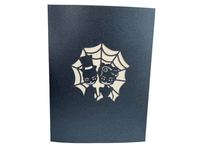 May include: A black card with a white cutout design of a couple dressed in Halloween costumes standing in front of a spider web.