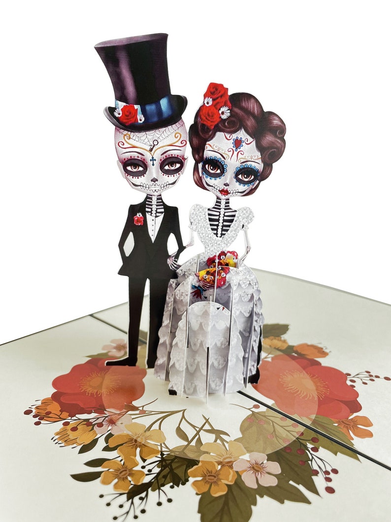 May include: A 3D pop-up card depicting a skeleton bride and groom in traditional Day of the Dead attire. The bride is wearing a white gown with lace detailing and a floral crown. The groom is wearing a black suit with a top hat. The card is decorated with a floral wreath.