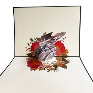 May include: A 3D pop-up card depicting a wedding ceremony with a floral wreath surrounding the couple. The couple are dressed in black and white, and the wreath is made of red, orange, and yellow flowers.