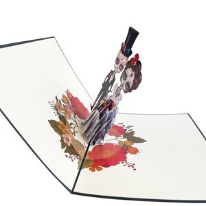 May include: A pop-up card depicting a skeleton couple dressed in wedding attire. The bride is wearing a white dress with red flowers and the groom is wearing a black suit with a top hat. The card is decorated with red and orange flowers.