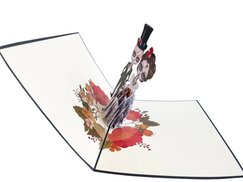 May include: A pop-up card depicting a skeleton couple dressed in wedding attire. The bride is wearing a white dress with red flowers and the groom is wearing a black suit with a top hat. The card is decorated with red and orange flowers.