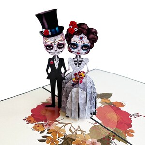 May include: A 3D pop-up card depicting a skeleton bride and groom in wedding attire. The bride is wearing a white gown with lace detailing and a red flower in her hair. The groom is wearing a black suit with a white shirt and a top hat. The card is decorated with colourful flowers.