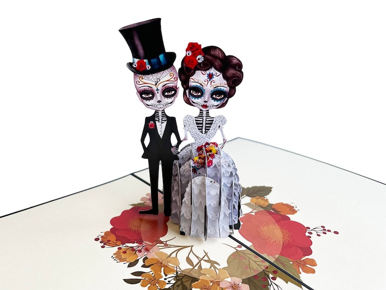 May include: A 3D pop-up card depicting a skeleton bride and groom in wedding attire. The bride is wearing a white gown with lace detailing and a red flower in her hair. The groom is wearing a black suit with a white shirt and a top hat. The card is decorated with colourful flowers.