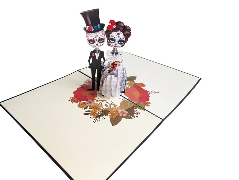 May include: A 3D pop-up card depicting a skeleton bride and groom in traditional wedding attire. The bride is wearing a white dress with floral embellishments and a red flower in her hair. The groom is wearing a black suit and a top hat. The card is adorned with a floral wreath.