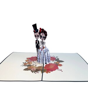 May include: A 3D pop-up card depicting a skeleton bride and groom in wedding attire. The bride is wearing a white dress with a floral pattern and a red flower in her hair. The groom is wearing a black suit and a top hat. The card is decorated with a floral wreath.