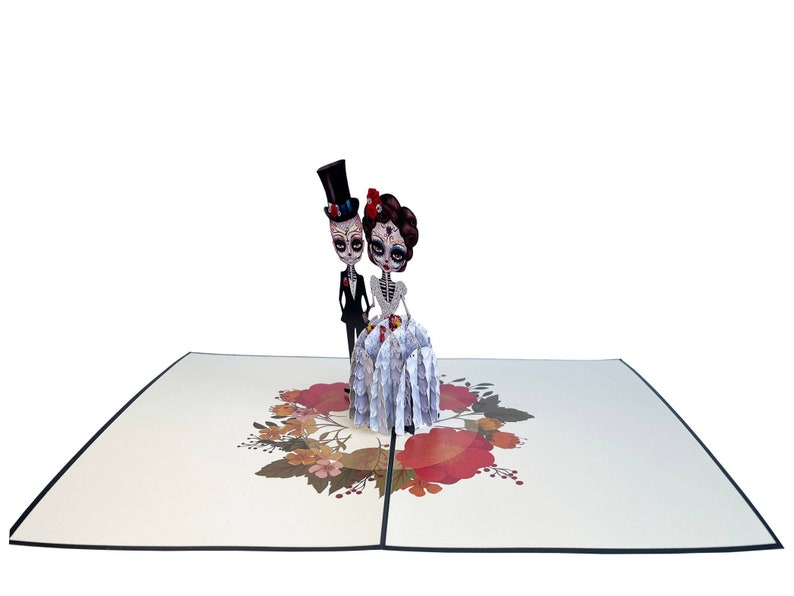 May include: A 3D pop-up card depicting a skeleton bride and groom in wedding attire. The bride is wearing a white dress with a floral pattern and a red flower in her hair. The groom is wearing a black suit and a top hat. The card is decorated with a floral wreath.