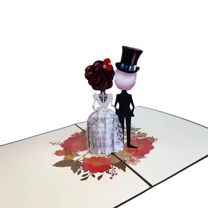 May include: A 3D pop-up card depicting a skeleton bride and groom in wedding attire. The bride is wearing a white gown with lace detailing and a red flower in her hair. The groom is wearing a black suit and a top hat. The card is decorated with red and orange flowers.