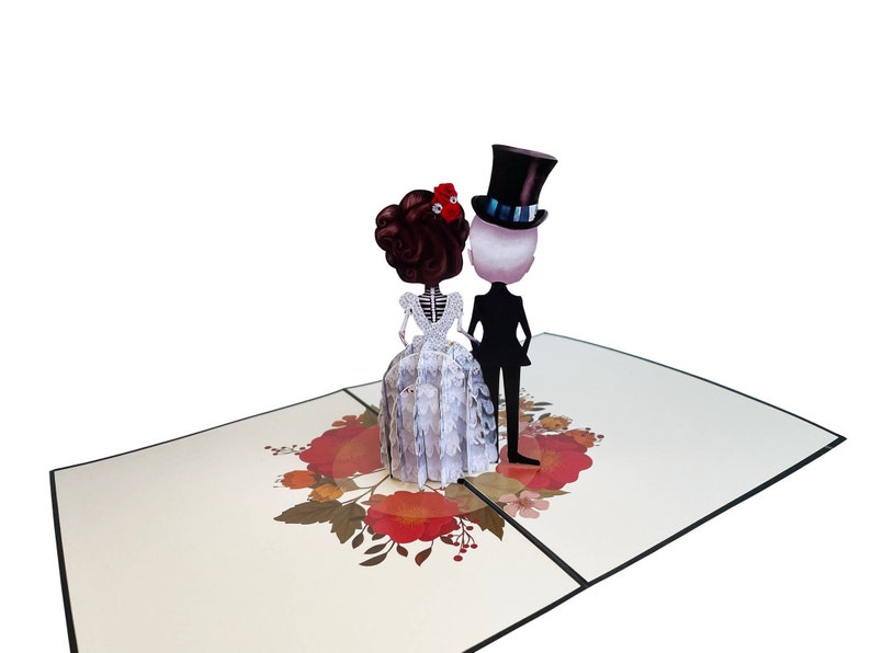 May include: A 3D pop-up card depicting a skeleton bride and groom in wedding attire. The bride is wearing a white gown with lace detailing and a red flower in her hair. The groom is wearing a black suit and a top hat. The card is decorated with red and orange flowers.
