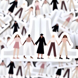 May include: Five women of different ethnicities and in different outfits stand holding hands in front of a white building. The women are wearing pink, white, black, brown, and beige clothing. The women are smiling and looking at the camera.