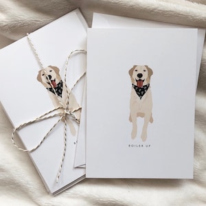 May include: Two white cards with a brown string tied around them. The card on top features a cartoon illustration of a yellow labrador retriever wearing a black bandana. The text "BOILER UP" is printed below the dog.