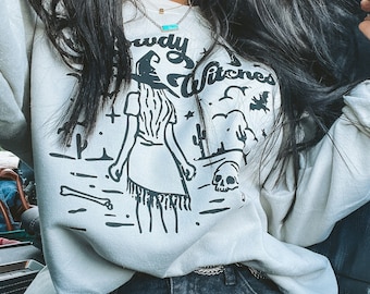 May include: A white sweatshirt featuring a black graphic of a witch wearing a cowboy hat and the text "Cowgirl Witches". The witch is depicted in a desert landscape with cacti and a skull. The sweatshirt is being worn by a person with long black hair and a turquoise necklace.