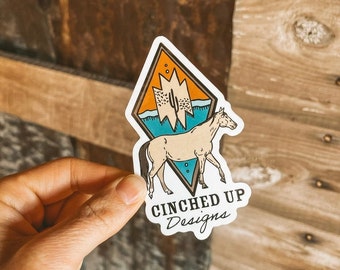 May include: A white diamond-shaped sticker featuring a brown and orange mountain scene, a blue body of water, and a white horse. The sticker has the text "CINCHED UP Designs" at the bottom.