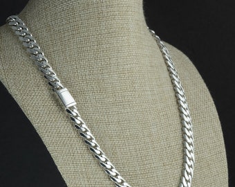 May include: A silver-colored curb chain necklace displayed on a mannequin. The necklace has a thick, interlocking link design and a rectangular clasp. The background is a dark color, and the mannequin is covered in a tan fabric.