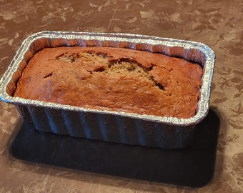 Può includere: Un filone di pane alla banana appena sfornato in una teglia usa e getta di alluminio. Il pane ha una crosta marrone dorata e si trova su una superficie nera. Lo sfondo è un motivo floreale marrone.