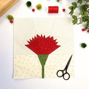 Pode incluir: Uma flor de cravo vermelho com caule e folhas verdes, feita de retalhos de tecido, é colocada sobre uma superfície branca com uma tesoura e outros materiais de artesanato.