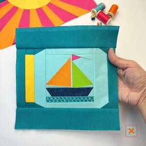 May include: A teal fabric square with a yellow, blue and green sailboat appliqué in the centre. The sailboat has a white mast and a pink sail. The fabric square is surrounded by a teal border.
