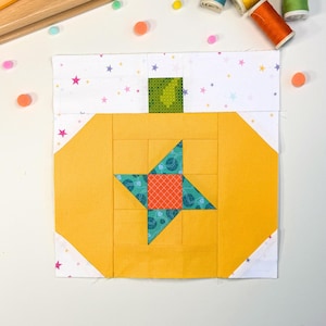 Pode incluir: Um bloco de colcha quadrado com um design de abóbora amarela com caule verde, um padrão de estrela central em azul-petróleo e laranja, e fundo branco com estrelas coloridas. Inclui um cortador rotativo amarelo, régua e quatro carretéis.