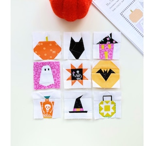 Pode incluir: Nove blocos de colcha quadrados de tecido com designs de Halloween, incluindo uma abóbora, gato preto, casa, fantasma, esqueleto, morcego, poção, chapéu de bruxa e lanterna, dispostos em uma grade com uma abóbora de feltro e um padrão.