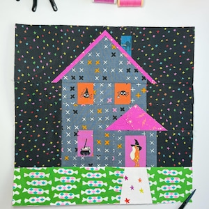 May include: A fabric quilt block featuring a house design with a black background printed with colorful stars. The house is gray with a cross-stitch pattern, a bright pink roof, and orange windows containing illustrations of a spider and a witch hat.