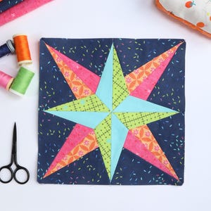 Pode incluir: Um bloco de colcha feito à mão com um desenho de estrela. A estrela é composta por tecidos rosa, laranja e verde sobre um fundo azul marinho. Materiais de costura, incluindo carretéis de linha e tesouras, são dispostos ao redor do bloco.