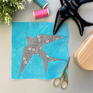 Pode incluir: Um quadrado de tecido azul turquesa com um desenho de andorinha de padrão cinza e branco. O tecido é costurado em estilo patchwork. Há também duas bobinas de fio, uma rosa e uma azul, e uma figura de andorinha de cerâmica preta.