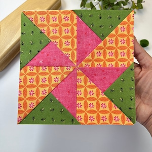 Puede incluir: Un bloque de colcha de retazos con un diseño de molinillo. El bloque está hecho de seis triángulos en colores alternos de verde, rosa y naranja con un patrón floral.