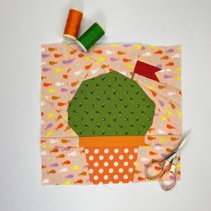 Pode incluir: Um bloco de tecido colorido com um desenho de cacto num vaso, com uma pequena bandeira vermelha. O bloco é rodeado por tecido rosa com um padrão de peixes. Também estão presentes carretéis de linha laranja e verde e pequenas tesouras.