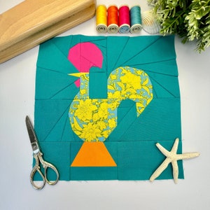 Pode incluir: Um bloco de colcha de retalhos turquesa com um design de galo. O galo é feito de tecido amarelo, rosa e branco com um padrão floral. O galo está em pé sobre uma base de tecido laranja.