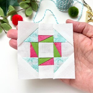 Pode incluir: Um pequeno quadrado de tecido branco com um design de patchwork verde e rosa. O design é uma estrela de quatro pontas com um quadrado no centro.