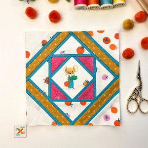 Op de afbeelding: Een wit vierkant van stof met een kleurrijk patchwork-ontwerp. In het midden is een cartoonmuis te zien die een bosje groene groenten vasthoudt. De stof is omgeven door een rand van oranje, blauwe en gele stroken.