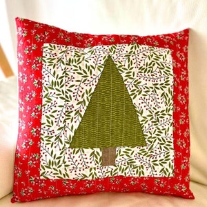 Könnte beinhalten: Ein rotes und grünes Weihnachtskissen mit einem gesteppten grünen Baumdesign in der Mitte. Der Baum ist von einem weißen Hintergrund mit einem Muster aus grünen Blättern und roten Beeren umgeben. Das Kissen hat einen roten Rand mit einem weißen Blumenmuster.