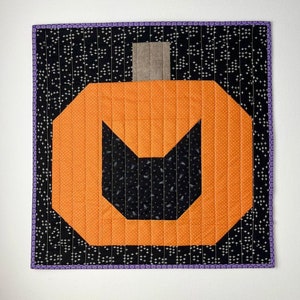 Pode incluir: Uma abóbora acolchoada preta e laranja com um recorte de rosto de gato preto. A abóbora é cercada por um fundo preto com bolinhas brancas e uma borda roxa.