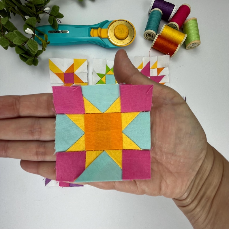 Peut inclure: Un bloc de patchwork carr&eacute; en tissu avec un carr&eacute; orange central entour&eacute; de triangles bleu clair et jaunes, et des carr&eacute;s roses aux coins. Le bloc est accompagn&eacute; d'un cutter rotatif turquoise et de plusieurs bobines de fil &agrave; coudre color&eacute;.