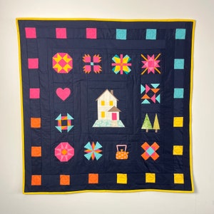 Pode incluir: Um cobertor azul marinho com uma borda amarela. O cobertor é feito de vários quadrados e formas coloridas, incluindo uma casa, um bule e árvores. O cobertor é perfeito para adicionar um toque de fantasia a qualquer cômodo.