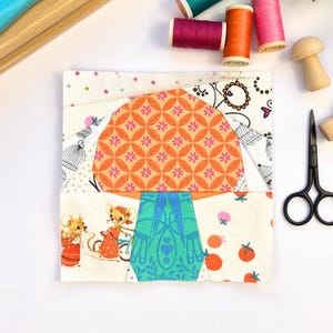 Pode incluir: Um cogumelo de tecido patchwork com um caule turquesa e uma tampa laranja com um padrão floral branco e rosa. O cogumelo é rodeado por quadrados de tecido com padrões diferentes, incluindo ratos, pássaros e flores.