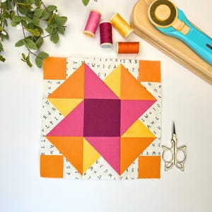 Pode incluir: Um bloco de colcha colorido com um design de estrela geométrica, apresentando triângulos laranja, rosa e amarelos e um quadrado central magenta escuro. O bloco é cercado por pequenos quadrados laranja e fica em um tecido branco com letras impressas. Carretéis de linha e um cortador rotativo também são visíveis.