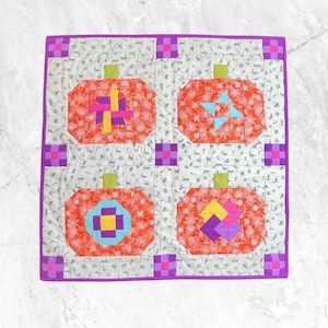 Puede incluir: Una colcha con ribete morado y cuatro bloques naranjas en forma de calabaza. Cada bloque presenta un diseño geométrico diferente en rosa, amarillo, azul y morado.