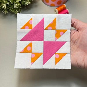 Pode incluir: Um bloco de patchwork feito à mão com um design geométrico. O bloco é composto por tecidos brancos, rosa e laranja. Os triângulos laranja têm bolinhas rosa. O bloco é segurado em uma mão.