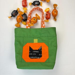 Pode incluir: Sacola de tecido verde com um bolso em forma de abóbora que apresenta uma silhueta de gato preto. A sacola tem alças de bolinhas brancas e está cheia de doces de Halloween.