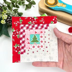 Pode incluir: Um bloco de colcha de retalhos vermelho, branco e rosa com uma árvore de Natal verde no centro. O bloco é feito de vários tecidos com padrões diferentes, incluindo xadrez, bolinhas e flocos de neve.
