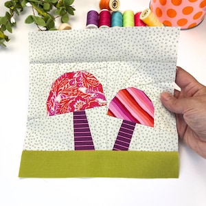 Pode incluir: Uma peça de arte têxtil feita à mão com dois desenhos de cogumelos. As tampas dos cogumelos são em tons de rosa e vermelho, com padrões listrados e florais. Os caules são roxos e a base é verde. O fundo é um tecido cinza claro com pequenos padrões de cruzes e círculos.