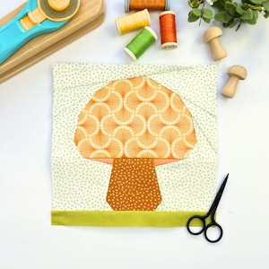 Pode incluir: Um aplique de tecido em forma de cogumelo com um esquema de cores verde, laranja e marrom. O cogumelo está sobre um fundo branco com uma borda verde.