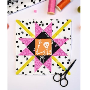 May include: A square fabric quilt block featuring a central orange square with a white skeleton graphic. The block is surrounded by a star pattern in pink floral and black fabric, set against a white background with black polka dots and small spiders.