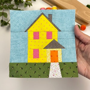 Pode incluir: Uma casa amarela com telhado marrom e uma porta branca. A casa tem duas janelas com cortinas rosa e um gramado verde com pequenas flores brancas. A casa está contra um céu azul.