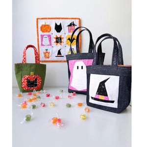 Pode incluir: Três sacolas de tecido com aplicações de Halloween: uma bolsa verde com uma abóbora, uma bolsa de jeans escuro com um fantasma branco e uma bolsa de jeans escuro com um chapéu de bruxa. Atrás, há uma tapeçaria quadrada acolchoada com nove ícones.