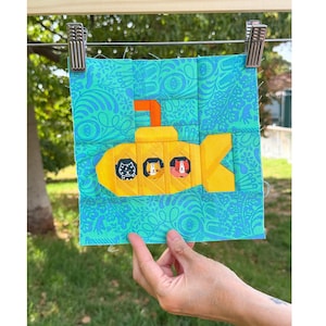 Può includere: Un blocco di tessuto quadrato per trapuntatura raffigurante un sottomarino giallo brillante con tre finestre circolari, ognuna contenente una diversa illustrazione di gatto. Lo sfondo è un tessuto verde acqua con un motivo floreale blu.