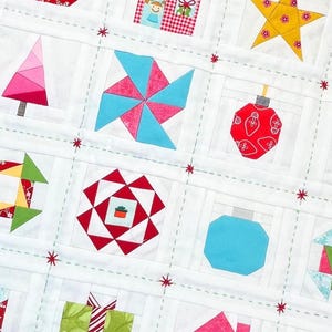 Pode incluir: Um edredão branco com vários desenhos temáticos de Natal. O edredão inclui quadrados com imagens de uma casa, estrela, árvore de Natal, enfeite e presente. Os desenhos são em vermelho, verde, amarelo e azul, com estrelas vermelhas.